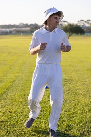 Young cricketer jogging in white uniform with bucket hat on sunny practice field