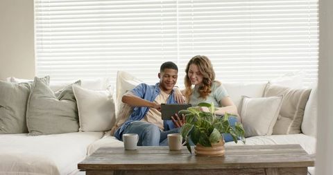 Diverse young couple relaxing on modern sectional sharing tablet with coffee and plants