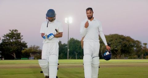 Two cricketers walking across pitch at dusk under floodlights carrying bats and helmets