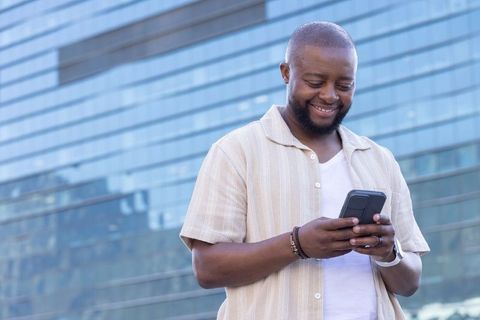Smiling man texting on smartphone in front of modern glass building, casual tech lifestyle