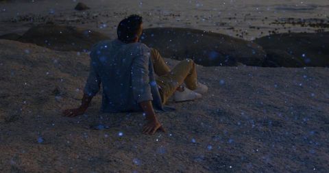 Solitary man sitting on rocky shore watching bioluminescent blue specks at dusk near boulders