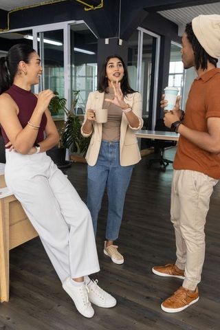 Diverse team chatting over coffee in open-plan office, woman gesturing while holding mug