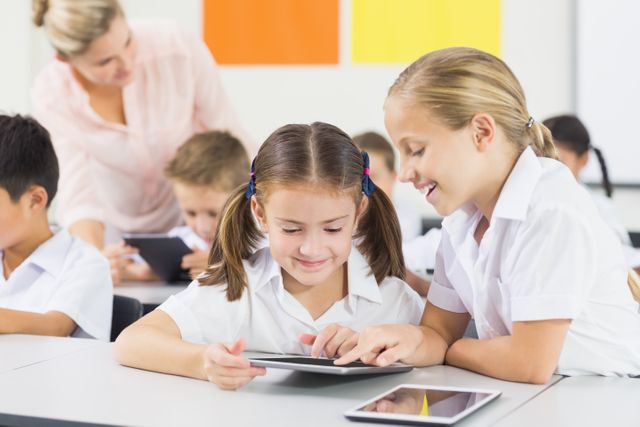 Young students in a classroom using digital tablets for learning activities. Teacher assisting in the background. Ideal for educational content, technology in education, modern classroom settings, and teamwork in learning environments.