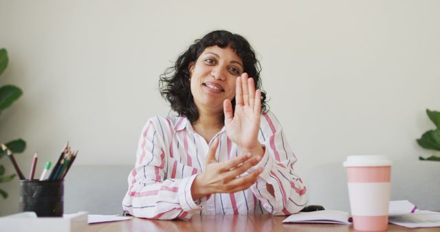 This image shows a smiling woman in a white blouse with red stripes and curly hair, gesturing with her hands during an office meeting. Papers, pens, and a coffee cup are on the desk. Ideal for use in websites, presentations, and marketing materials that focus on professional communication, office environments, and workplace collaboration.