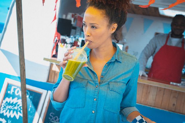Biracial woman in a denim shirt enjoying a healthy green drink at a food truck. Ideal for use in articles or advertisements about street food, small businesses, healthy lifestyles, and outdoor dining experiences. Can also be used to promote food trucks, catering services, and urban lifestyle content.