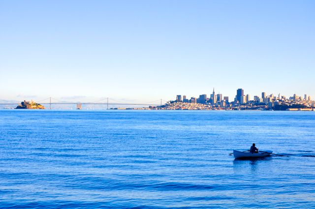 Person rowing small boat on calm water with city skyline in background. Ideal for use in travel and tourism promotions, highlighting serene outdoor activities, representing cityscapes, or emphasizing solitude and peacefulness in urban settings.