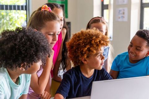 Multiracial elementary students collaborating around laptop in bright classroom