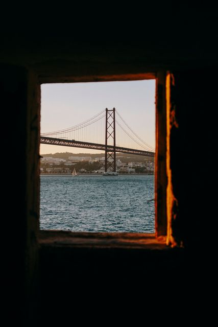 Capturing a stunning view of a bridge during sunset through a rugged window frame, with calm waters and a sailing boat. Ideal for travel advertisements, tourism websites, or inspirational posters. The image highlights the scenic charm and architectural beauty of coastal areas.