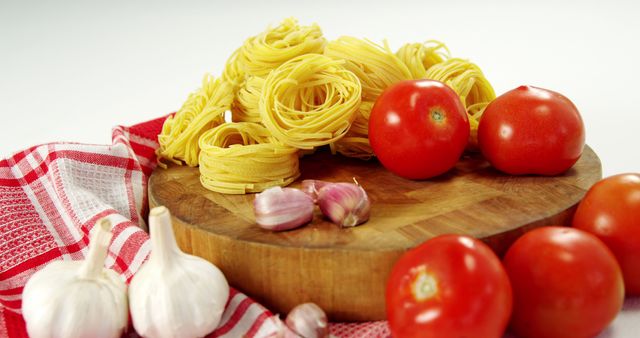 Fresh ingredients for cooking Italian pasta placed on a round wooden board. Yellow pasta nests are arranged alongside ripe tomatoes and whole garlic bulbs, accompanied by a red and white striped kitchen towel. Perfect for culinary blogs, recipe articles, and advertisements for Italian food products or cooking tutorials.