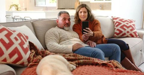 Couple Relaxing on Sofa Taking Selfie with Dog amid Cozy Knit Throws and Patterned Pillows
