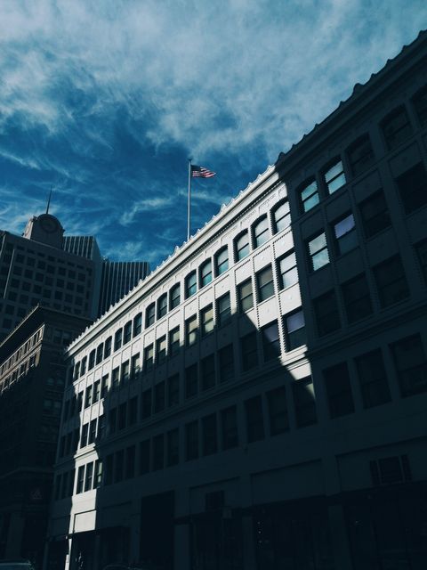 This image showcases urban buildings under a blue sky with shadows cast across the structures and a flag on one of the rooftops. Ideal for themes related to metropolitan living, architecture, city development, or patriotic elements when highlighting the flag.