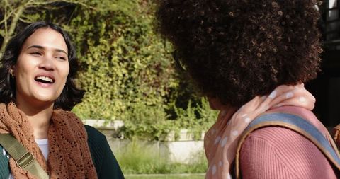 Two friends chatting outdoors wearing scarves and backpacks in sunny courtyard