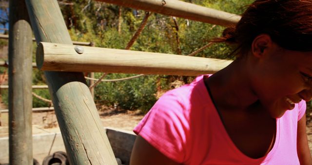 The image features a woman exercising outdoors on a wooden structure in a sunny environment. She is smiling and appears to be enjoying her workout. This image could be used in materials promoting healthy lifestyles, fitness routines, outdoor activities, and wellness programs.