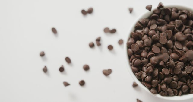 Close-up view of a white bowl filled with chocolate chips with some scattered around. This delectable image is perfect for use in food blogs, baking websites, or dessert recipe articles. It evokes a sense of indulgence and is ideal for advertising sweet products or culinary ingredients.