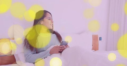 Asian woman relaxing in bed holding smartphone with cozy bokeh lighting and soft blanket