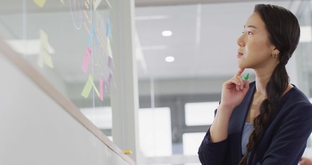 Businesswoman brainstorming on a clear glass board covered with colorful sticky notes in a modern office. Ideal for images featuring planning processes, corporate presentations, professional environments, strategic thinking, and entrepreneurial activities. Can be used for business-related articles, workplace productivity discussions, and creativity representation.