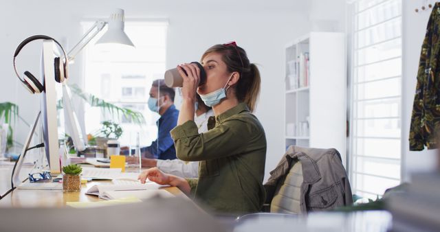 Woman seated at office desk, wearing face mask, taking a sip of coffee. Colleague in background working on computer. Office setting with modern equipment and decor. Can be used in workplace safety guidelines, remote work setup, health in workplace, or corporate communication materials.