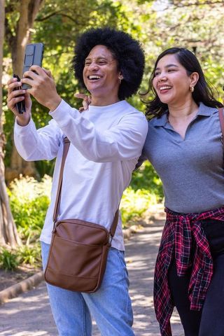Diverse couple smiling and taking selfie while walking through sunny park greenery