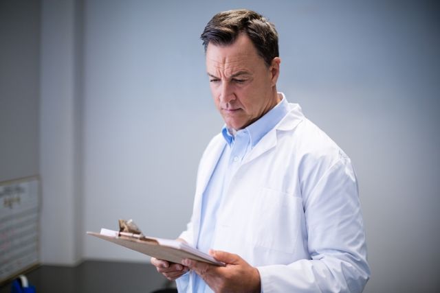 Male doctor in white coat reading reports on clipboard in hospital corridor. Ideal for use in healthcare, medical, and hospital-related content, including articles, brochures, and websites focused on patient care, medical staff, and clinical settings.