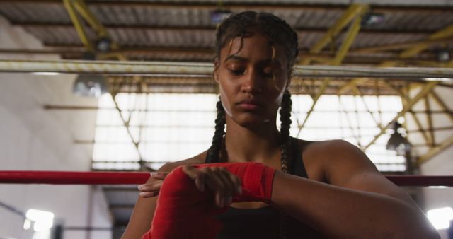 This image captures a focused biracial female boxer with braided hair wrapping her hands as she prepares for a training session. The photo emphasizes her determination, strength, and commitment to her sport. This stock photo can be used in articles and content related to boxing, sports training, women's empowerment, athletic preparation, and mental focus. Great for use on fitness blogs, motivational content, sports advertisements, and websites promoting physical activity and training programs.