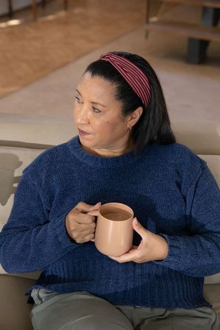 Mature woman holding ceramic mug on sofa in cozy living room, wearing navy sweater and red headband