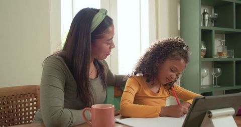 Mother guiding daughter with homework at tablet on wooden dining table, online learning