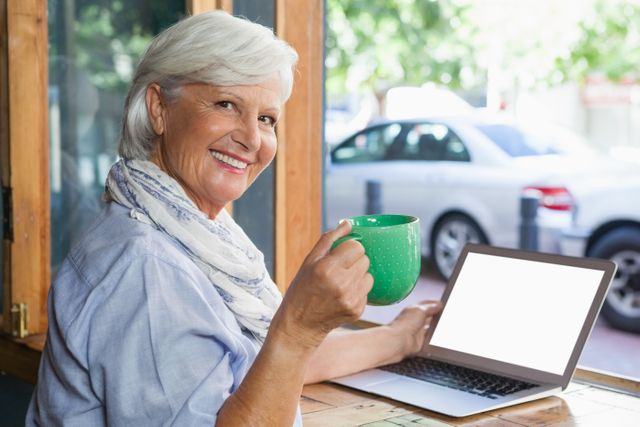 Senior woman enjoying coffee while using laptop in a cafe. Ideal for themes related to senior lifestyle, technology use among elderly, relaxation, and casual dining. Perfect for advertisements, blogs, and articles focusing on active aging, digital literacy, and social activities for seniors.