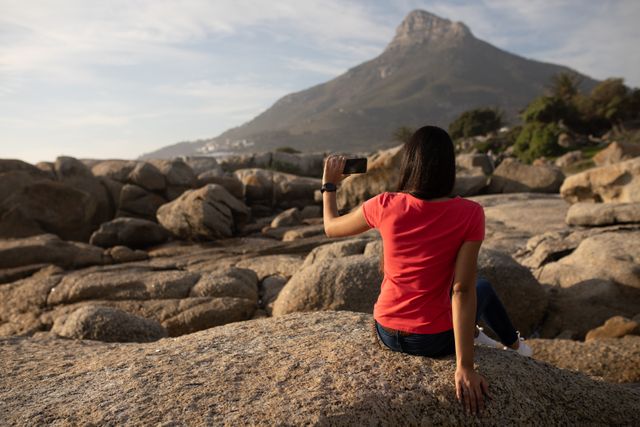 Rear view of a biracial woman enjoying free time in nature on a sunny day, sitting on a rock, admiring the view, holding a smartphone, taking photo of the countryside. 