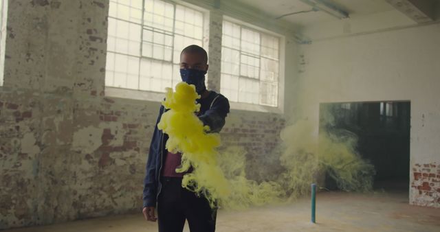 Young biracial man holds a smoke bomb indoors, with copy space. Vivid yellow smoke adds a dynamic effect to the industrial setting.