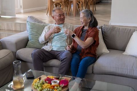Asian senior couple toasting with cups on cozy gray sofa, fruit platter on table