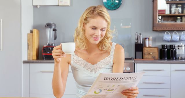 Young blonde woman smiling while reading a newspaper and drinking coffee in her home's kitchen. Perfect for illustrating concepts related to morning routines, home lifestyle, relaxation, or casual reading. Suitable for articles, blog posts, and advertisements focusing on domestic life, personal time, and everyday comfort.