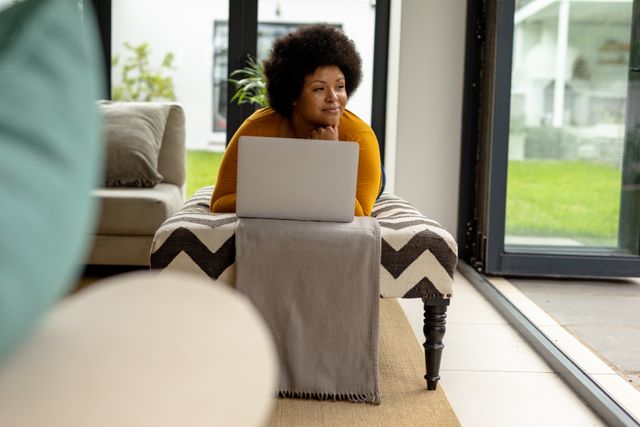 Smiling thoughtful african american mid adult woman looking away while using laptop at home. unaltered, lifestyle, wireless technology, contemplation, relaxation and domestic life concept.