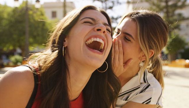 Two friends laughing and whispering in sunlit urban plaza