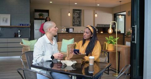 Two diverse women discussing over coffee at modern cafe table with smartphone and takeaway