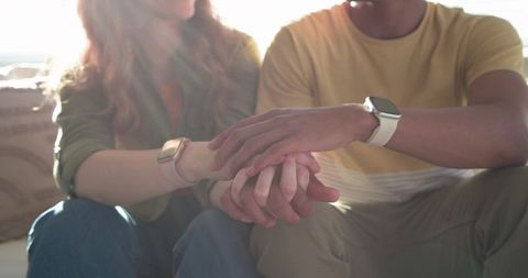 Sunlit multiracial couple holding hands on sofa with matching smartwatches and warmth