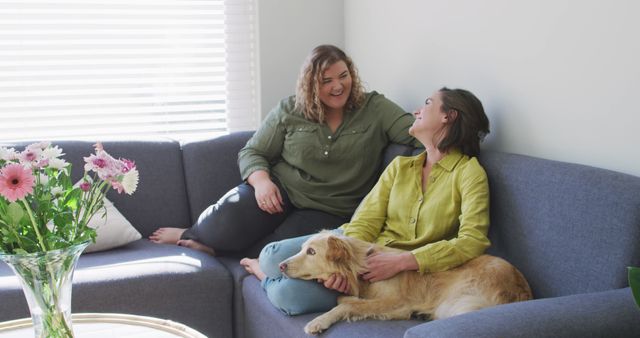 Women are sitting comfortably on a modern sofa, chatting and enjoying each other's company while a dog rests on one's lap in an inviting living room. Fresh flowers in a clear vase add a homely touch. Ideal for use in promotions for friendship, home decor, pet companionship, and everyday joy.