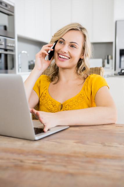 Smiling woman talking on mobile phone while using laptop in kitchen at home. Ideal for illustrating remote work, modern communication, multitasking, and domestic life. Suitable for articles, blogs, and advertisements related to home office, technology, and work-life balance.