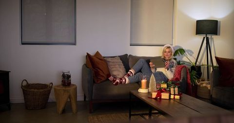 Cozy holiday evening with woman relaxing on sofa, using tablet amid candles and gifts
