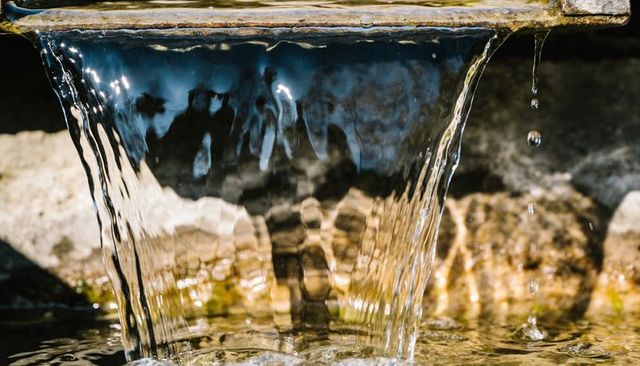 Sunlit water cascading from fountain lip flowing into shallow stone basin with droplets