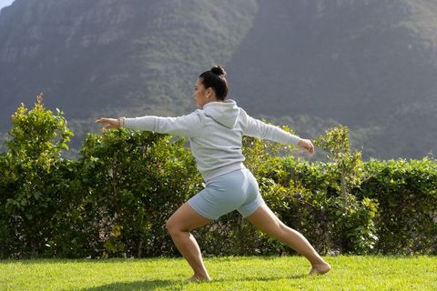 Woman practicing warrior lunge on green lawn with mountain backdrop wearing hoodie