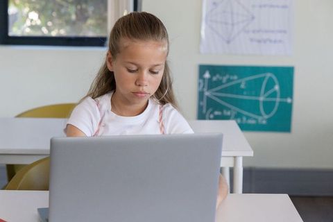 Preteen girl studying on laptop in bright classroom with geometry chart and focus