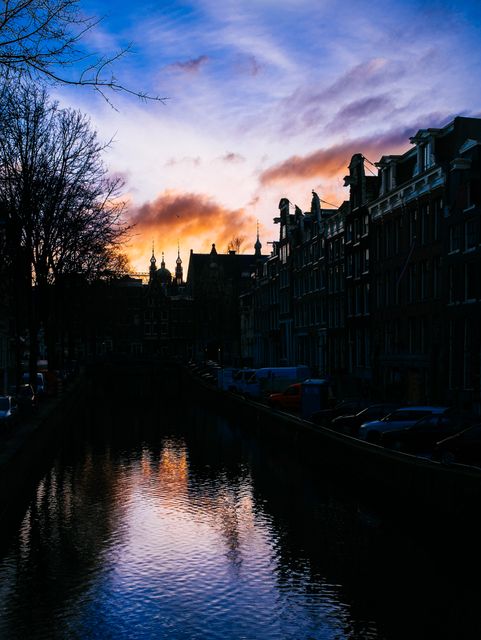 Amsterdam's iconic canal reflecting vibrant sunset colors, creating a tranquil and serene atmosphere. Ideal for illustrating peaceful cityscapes, travel advertisements, tourism promotions, and serene evening settings.