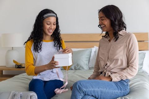 Teen daughter opening gift box with smiling mother on bed, warm family bonding