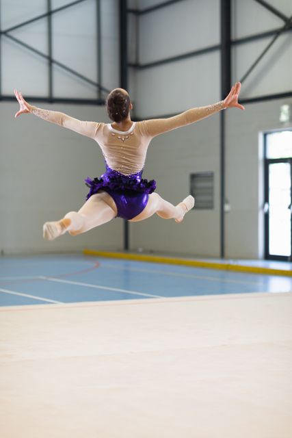 Rear view of a Caucasian female gymnast practicing at the gym, jumping and doing splits in the air with her arms outstretched. Gymnast training hard for competition.