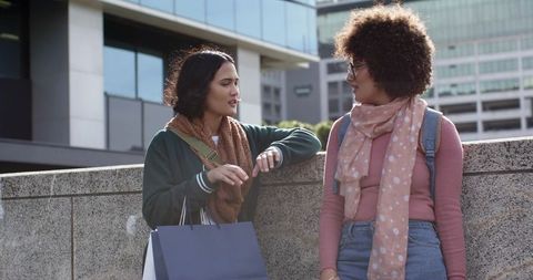 Two friends leaning on concrete ledge, chatting in sunlit urban plaza with shopping bag