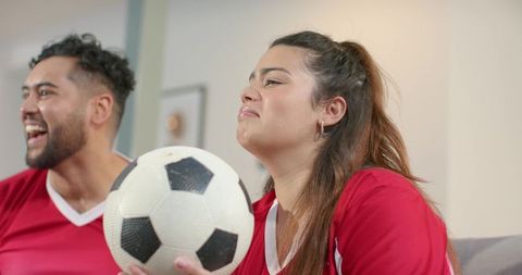 Tense soccer fans on couch holding ball, cheering and expecting game moment in red jerseys