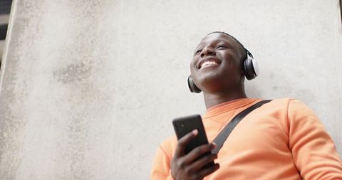 Smiling man listening to music on smartphone with headphones, leaning on concrete wall
