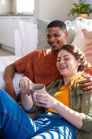 Diverse couple lounging on sofa sharing coffee and laughter in sunlit modern apartment