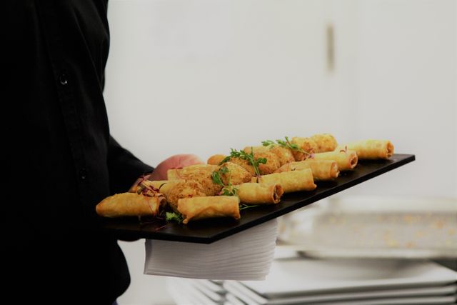 Waiter holding tray filled with freshly prepared appetizers including spring rolls and other delicacies, ideal for event planning, catering services, promoting hospitality industry, showcasing food presentation skills, and menus for social gatherings or corporate functions.