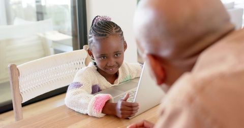 Adult male leaning toward african american girl sharing laptop and smiling at home dining table